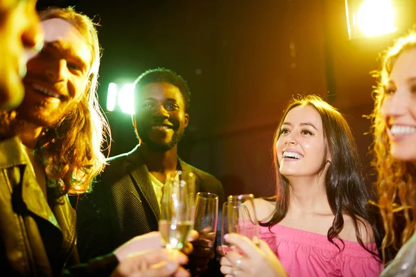 Group of positive excited young multiethnic friends standing in nightclub with colorful lights and clinking flutes while talking at party