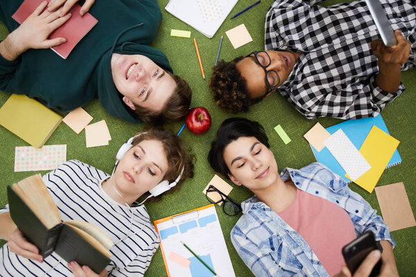 Высокий угол обзора расслабленных современных мультиэтнических студентов в повседневной одежде, лежащих в кругу на полу с грязными наклейками и рабочими книгами при использовании гаджетов и чтении книг
.