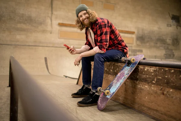 Uzun saçlı patencinin akıllı telefon kullanarak ve aşırı parkta rampada otururken kameraya gülümseyen tam uzunlukta portre, kopyalama alanı