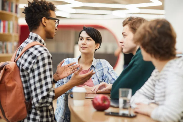 Group of content multiethnic high school student friends in casual clothing standing at counter in library and discussing project