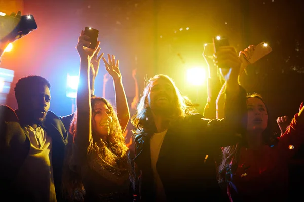 Jolly energetic young people standing in projector light and waving hands with smartphones while screaming at musical concert