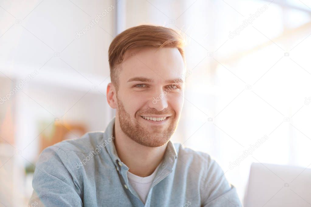 Head and shoulders portrait of handsome young man looking at camera  sitting at workplace in office and smiling , copy space