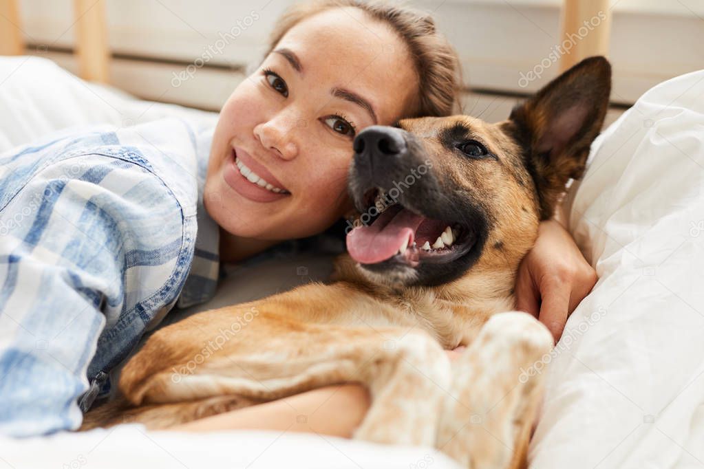 Portrait of smiling Asian woman hugging dog lying on bed  together and looking at camera, copy space