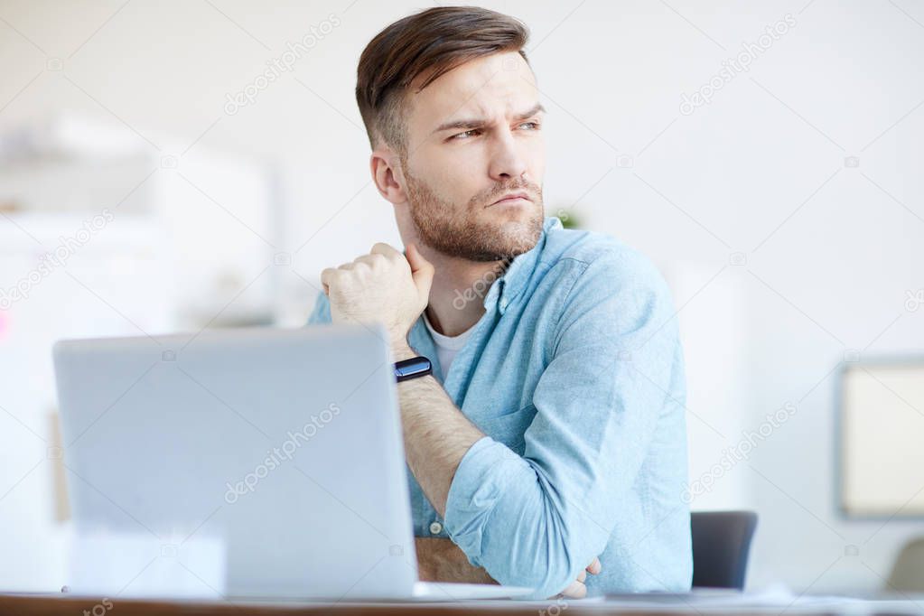 Portrait of handsome businessman looking away pensively while sitting at workplace in office, copy space