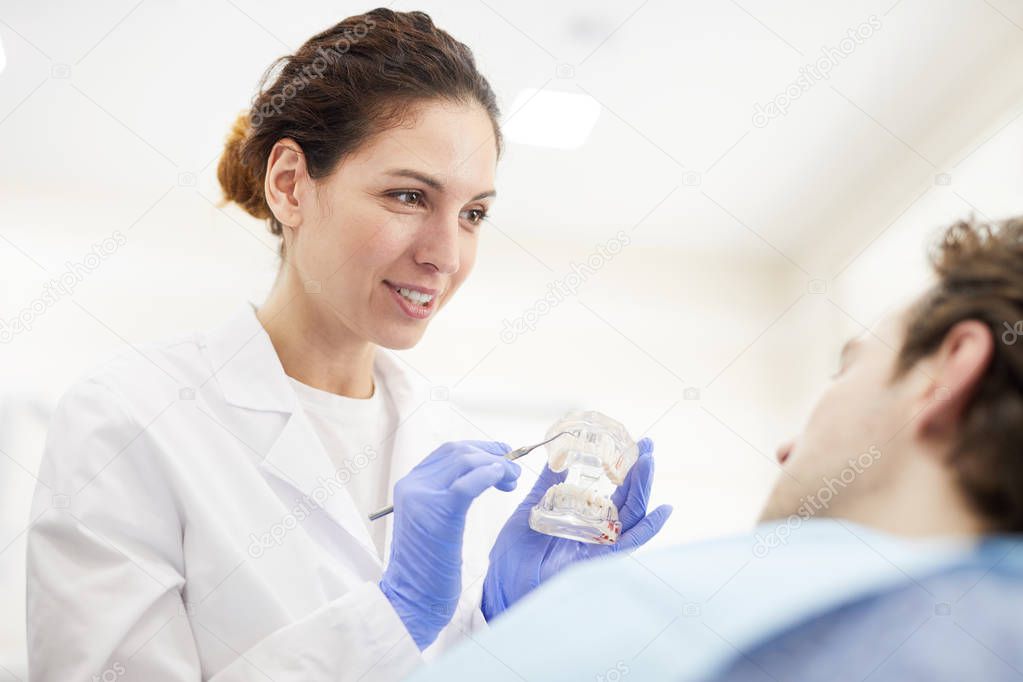 Portrait of female dentist showing tooth model to patient sitting in dental chair, copy space