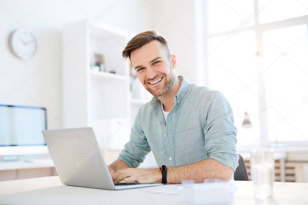Portrait of handsome young man smiling at camera while using laptop in office, copy space