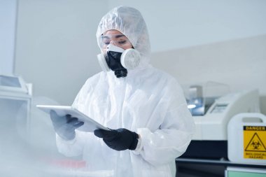 Testlerin sonuçlarını incelerken dijital tablet kullanarak koruyucu kostüm ve solunum maskesi meşgul laboratuvar biyolog
