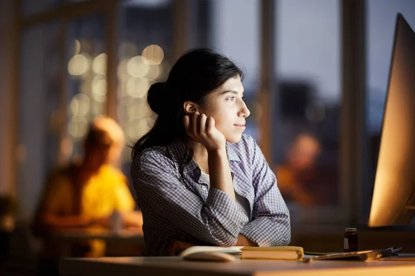 Side view portrait of dreamy young woman looking at computer screen while working late in dark office, copy space