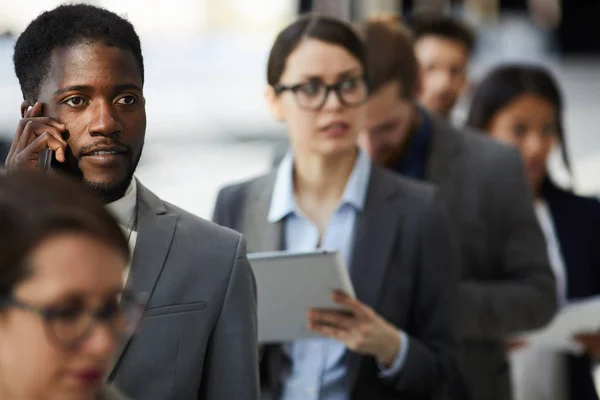 Business forum participants waiting for registration in line: serious handsome African-American businessman with beard talking on pone and looking at big queue in front of him