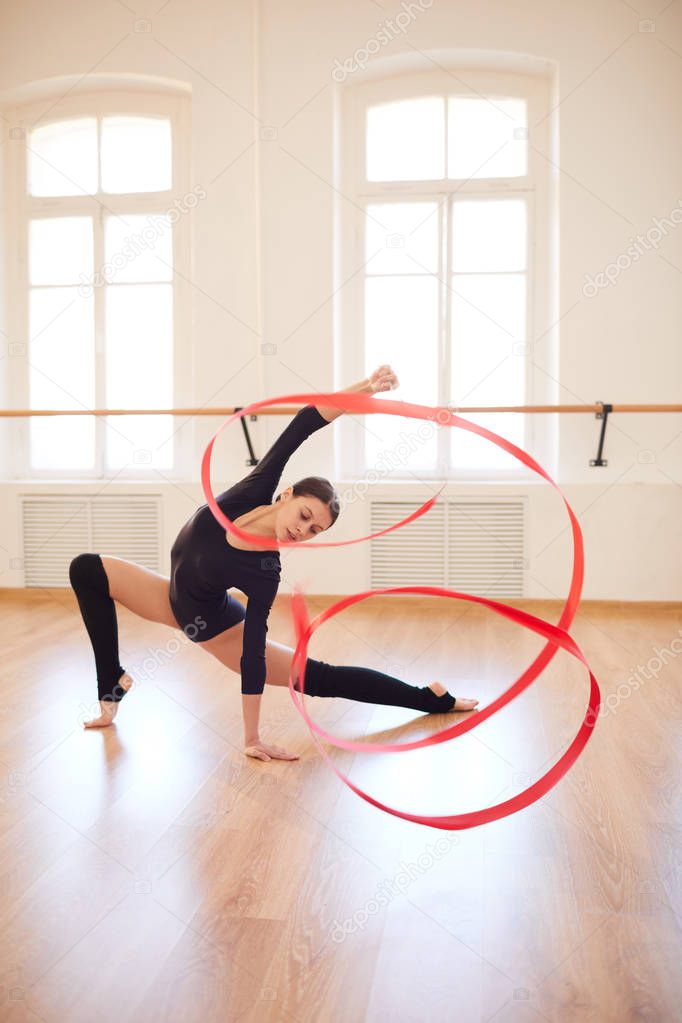 Slim artistic pretty girl in tight bodysuit leaning on floor and making circle with ribbon while dancing with gymnastic ribbon in empty studio
