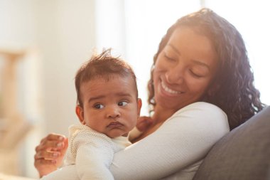 Kanepede oturan ve evde kollarda bebek tutan mutlu heyecanlı kıvırcık saçlı Genç Afrikalı kadın 