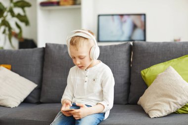 İçerik meraklı teknoloji-küçük çocuk kulaklık kanepede oturan ve telefonda oyun oynamak bağımlısı