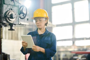 Atölyede makinelerin yanında duran tablet kullanarak hardhat takan kadın işçinin bel kadar portresi, kopyalama alanı