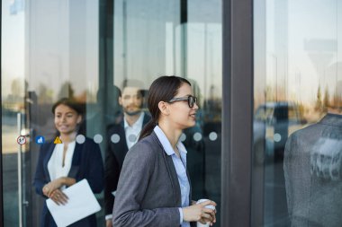 Kahve fincanı tutan ve ofis binasına gelirken döner kapıdan yürüyen gözlüklü gülümseyen çekici genç iş kadını
