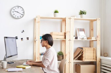 Ev ofisinde masada oturan bilgisayar kullanarak genç etnik kadının yan görünüm portresi, kopya alanı