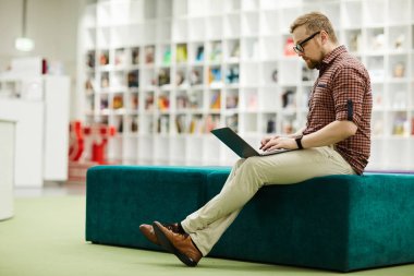 Rahat puf üzerinde oturan ve kütüphane alanında dizüstü bilgisayar ile çalışan rahat kıyafet ve gözlük Ciddi modern adam