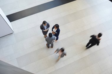 Şirket lobisinde duran ve mola sırasında sohbet eden formalgiyimli kültürlerarası iş arkadaşlarının görüşü doğrudan üzerinde, diğer çalışanlar acele ederken, bulanık hareket etkisi