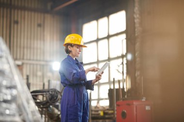 Endüstriyel atölyede duran dijital tablet kullanarak hardhat giyen güçlü kadının yan görünüm portresi, kopyalama alanı