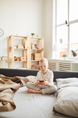 Ciddi Gadget bağımlısı çocuk koltuk yatak üzerinde çapraz bacaklar ile oturan ve evde net sörf yaparken modern cihaz kullanarak