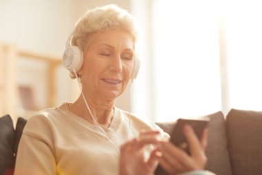 Güneş ışığı ile aydınlatılmış evde kanepede otururken akıllı telefon kullanarak müzik dinlerken gülümseyen kıdemli kadın portresi, kopyalama alanı