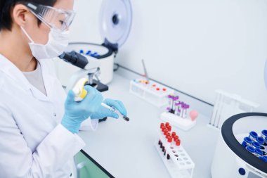 Beyaz önlük ve kauçuk eldiven ciddi meşgul genç kadın laboratuvar işçisi masada duran ve numuneler ile çalışırken test tüpükan ekleme