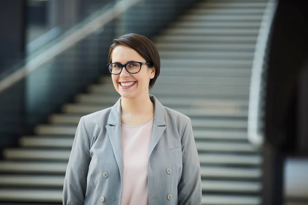 Cheerful excited female project manager with wide smile wearing gray jacket and eyeglasses standing in lobby and looking at camera