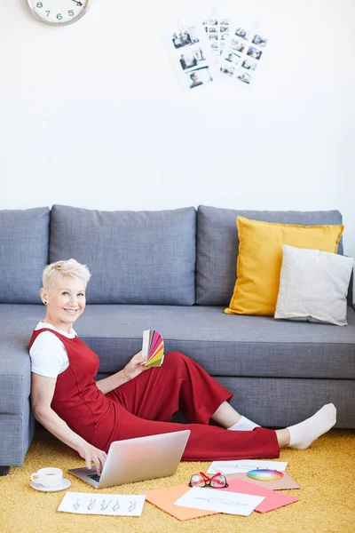 Young successful blonde creative woman in casualwear sitting on the floor by sofa while working with color swatches and fashion model sketches