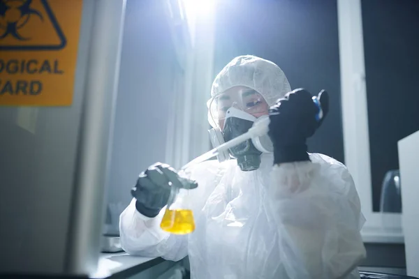 在实验室研究物质时 佩戴防护镜 生物危害套装和呼吸面罩的浓缩微生物学家将试剂放入烧瓶中 — 图库照片