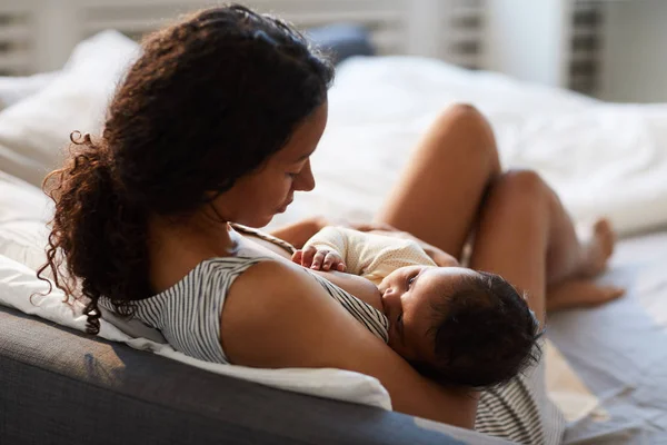 ベッドに座って 乳房で赤ちゃんを養っている間 ヘッドボードに寄りかかっている平和な愛情の若いアフリカの母親 — ストック写真