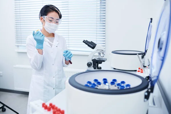 严重集中的年轻亚洲实验室研究员 短发穿着白色外套 口罩和安全护目镜混合试剂与血液样本在工作场所 — 图库照片