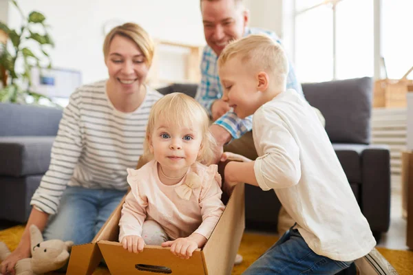 新しいフラットで楽しんでいる2人の子供を持つ肯定的な遊び心のある若い家族 動く箱に座って 他の家族がこの箱の中を見ている間 カメラを見ているかわいい女の子 — ストック写真