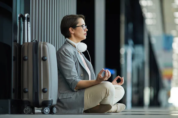 Serious concentrated middle-aged lady with headphones on neck sitting with crossed legs and holding hands in mudra while meditating before flight