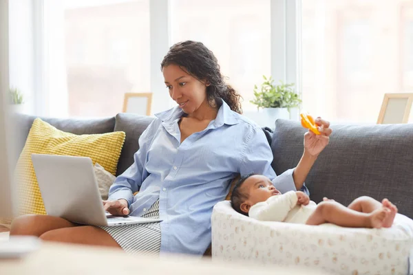 コンテンツは ソファに座って 仕事のためにラップトップを使用しながら コクーンで赤ちゃんの息子と遊ぶカジュアルな服を着て若い黒人の母親を集中 — ストック写真