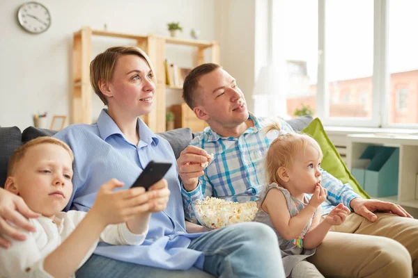 リビングルームのソファに座り ポップコーンを食べながらテレビを見ている好奇心旺盛な若い家族は テレビの前で余暇を過ごす — ストック写真