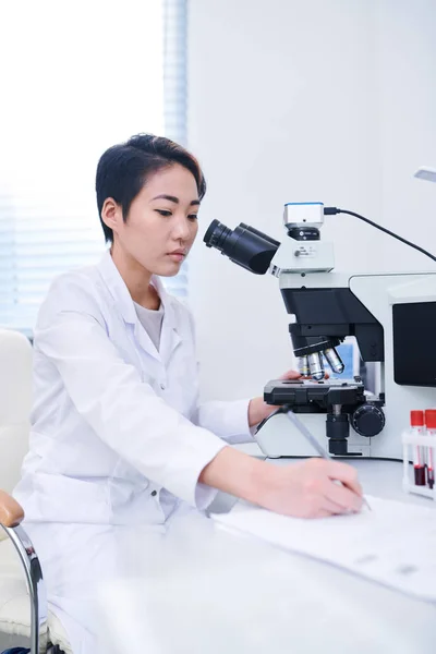 현미경으로 세포를 공부하는 테이블에 데이터를 코트에 심각한 아시아 생물학자 — 스톡 사진