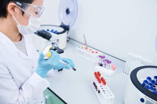 严重忙碌的年轻女实验室工作人员穿着白色外套和橡胶手套站在办公桌 并在试管中添加血液 同时处理样品 — 图库照片
