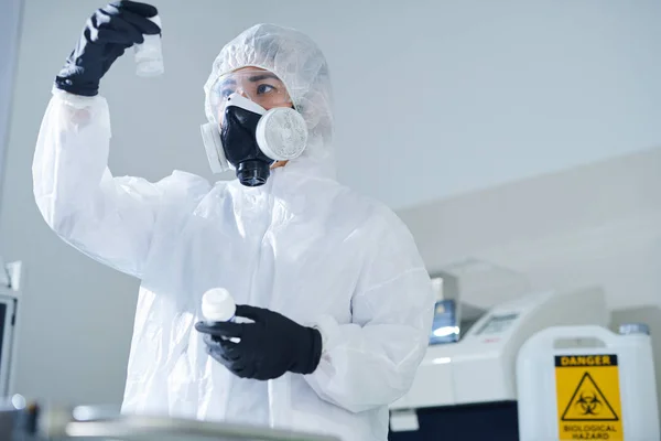 身着生物危害服和呼吸器的严重周到的有毒实验室工作人员站在隔离室 在实验室研究治疗药物时检查物质瓶 — 图库照片