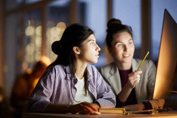 Portrait of two young women pointing at computer screen while working in dark office at night, copy space