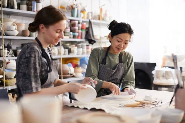 Positive attractive young multi-ethnic women in aprons sitting at table with craft tools and processing dishes with sandpapers in workshop — Stock Image Positive Attractive Young Multi Ethnic Women Aprons Sitting Table Craft Royalty Free Stock Images