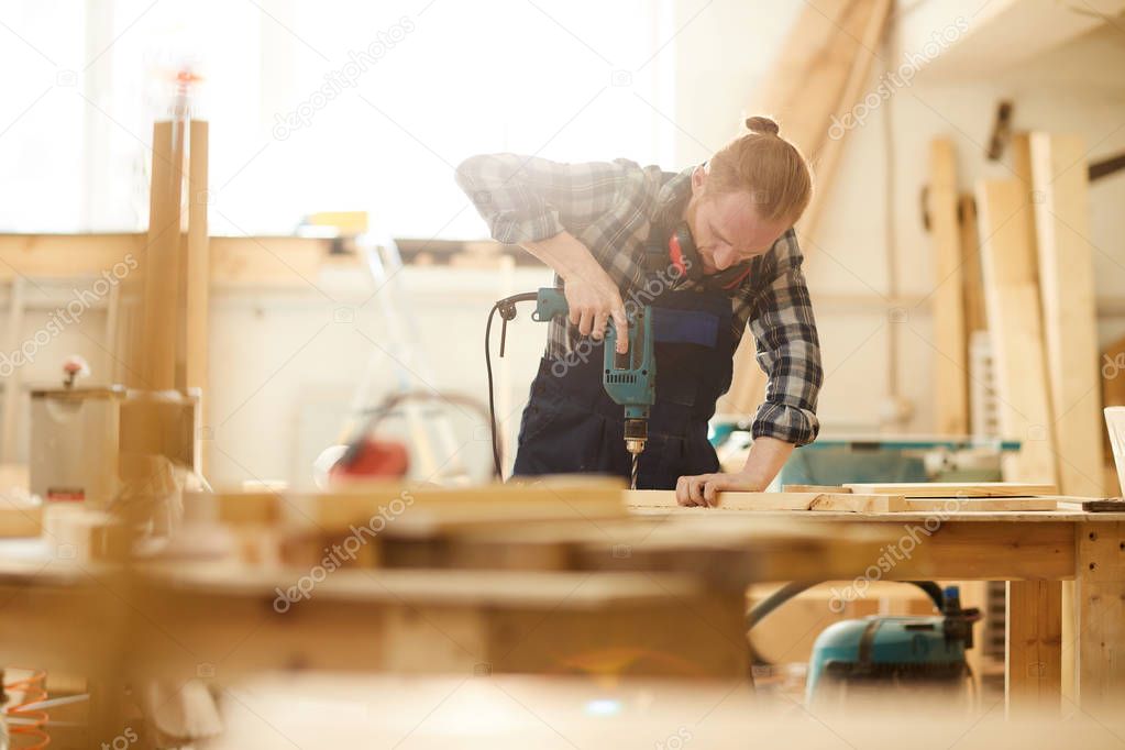 Waist up portrait of red haired carpenter drilling wood while working in joinery lit by sunlight, copy space