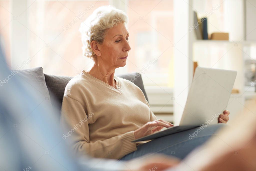 Warm toned portrait of contemporary mature woman using laptop sitting on sofa at home, copy space