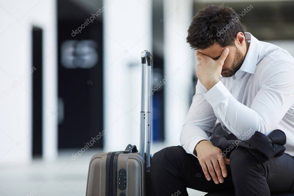 Sad handsome young businessman in white shirt sitting with wheeled luggage in airport and crying because of being late on plane