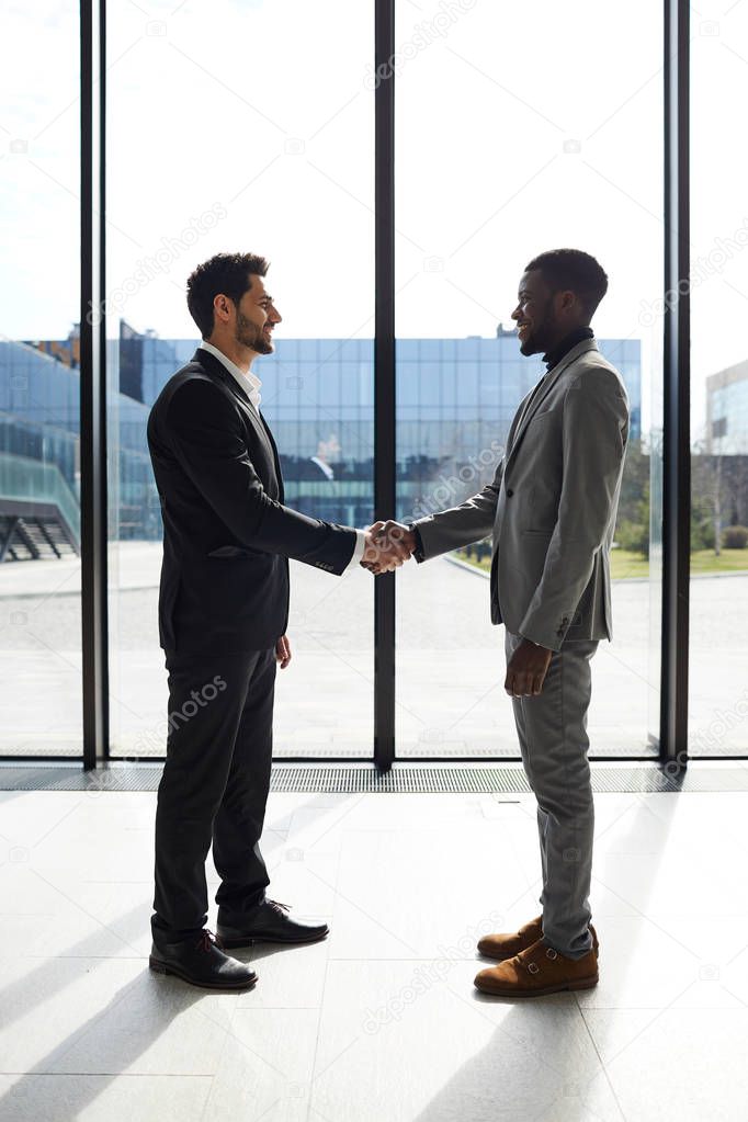 Cheerful confident young mixed race manager in black suit welcoming business partner to company and shaking hands with him in lobby