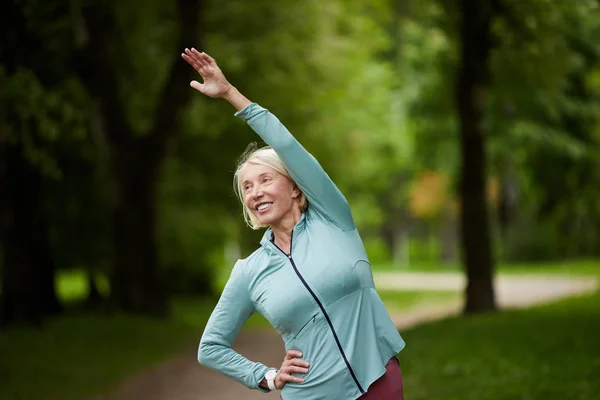 快乐金发活跃妇女在运动服做侧弯在公园绿色树木的背景锻炼 — 图库照片