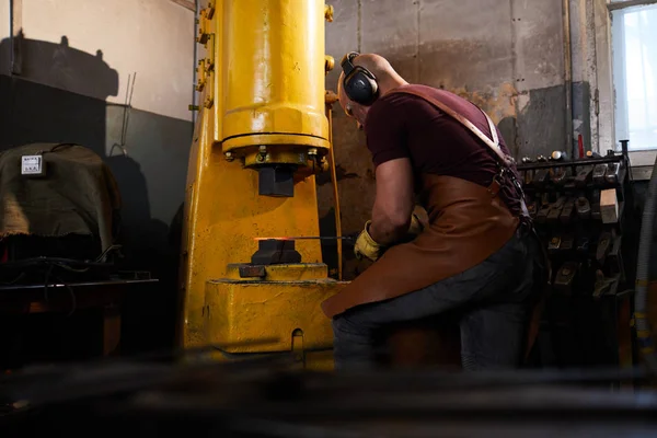 Concentrated skilled man in apron and ear protectors putting metal bar under press while working with hydraulic forging press