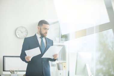 Ciddi meşgul genç sakallı işadamı takım elbise modern ofiste ayakta ve iş belgeleri ile çalışıyor