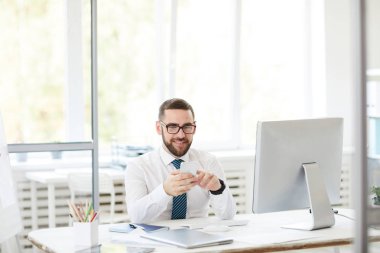 Ofiste çalışırken masada oturan ve akıllı telefon kullanarak gözlük gülümseyen yakışıklı genç sakallı yönetici