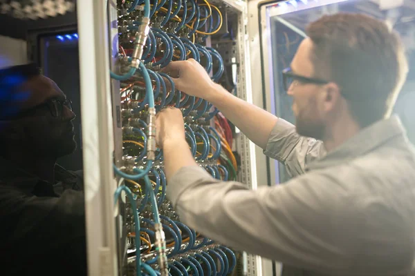 Busy young network engineer standing at open cabinet of mainframe and adjusting cables while fixing server