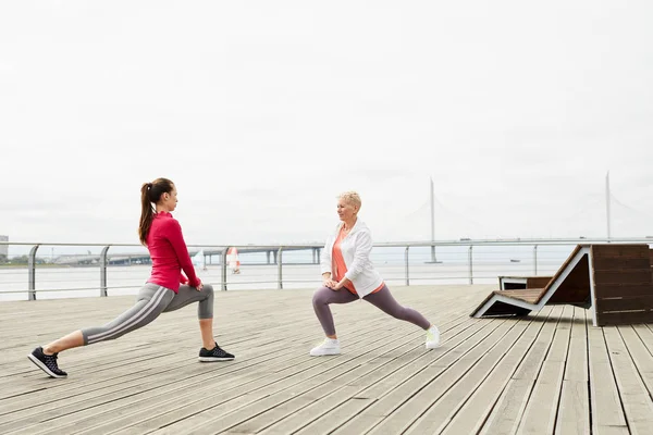 Side view portrait of two active young women stretching legs while doing yoga on wooden pier, copy space
