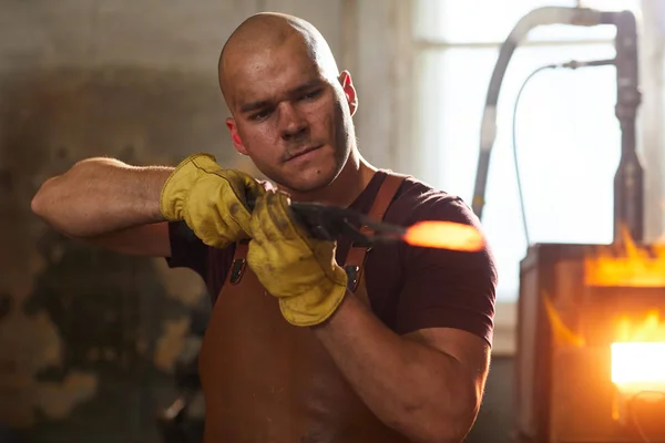 Concentrated young metalworker in apron and gloves holding heated metal piece in tongs while examining its shape